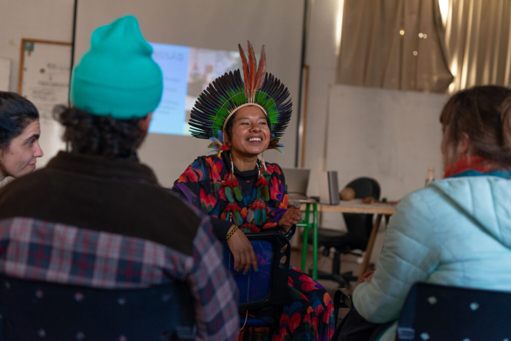 Mirna sentada en el salón sonriendo y con personas escuchándola.
