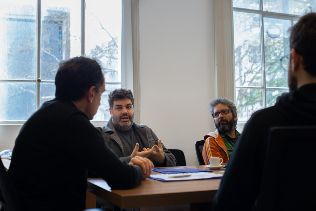 Fotografía donde se ve alrededor de una mesa al decano de Facultad de Artes Fernando MIranda de espalda y al director del Programa Apex MIguel Olivetti y Pablo Pereira.