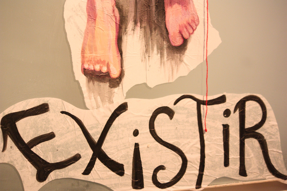 obra en papel adherido a la pared donde se ve detalle de pies y la palabra "existir"