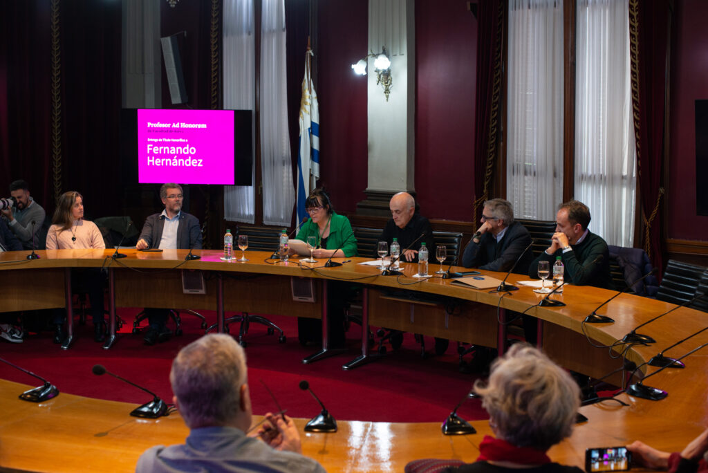 Sala Maggiolo de la Udelar donde se ven a las personas participantes sentadas alrededor de la mesa. Se ve haciendo uso de la palabra a Arianna Fasanello. 