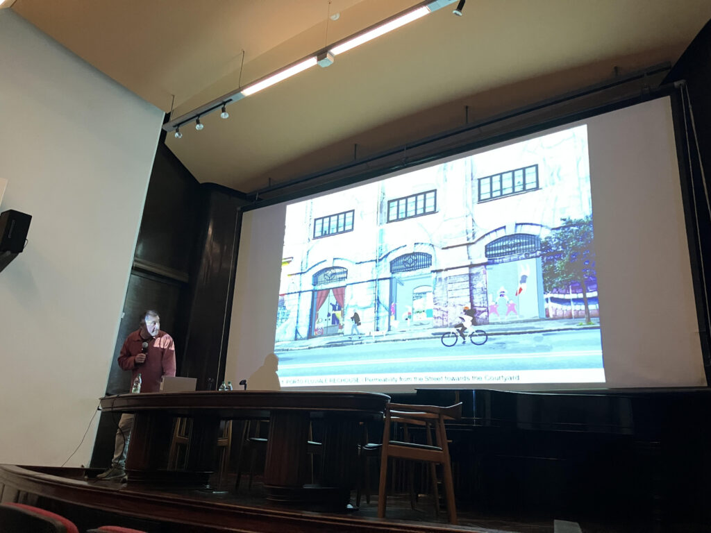 Fotografía de Francesco Careri durante la conferencia que brindó en el salón de actos de FADU. Se lo ve de pie pasando diapositivas al costado de la mesa de la sala y ante una pantalla gigante donde se ve la imagen de una vereda y calle de una ciudad, peatones y persona andando en bicicleta.