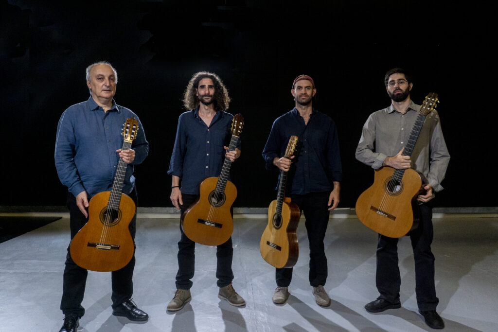 Fotografía de cuerpo entero de los integrantes de Eúm Cuarteto de Guittaras, con los cuatro integrantes que lo conforman donde se los ve sobre fondo negro sosteniendo cada uno de ellos una guitarra.