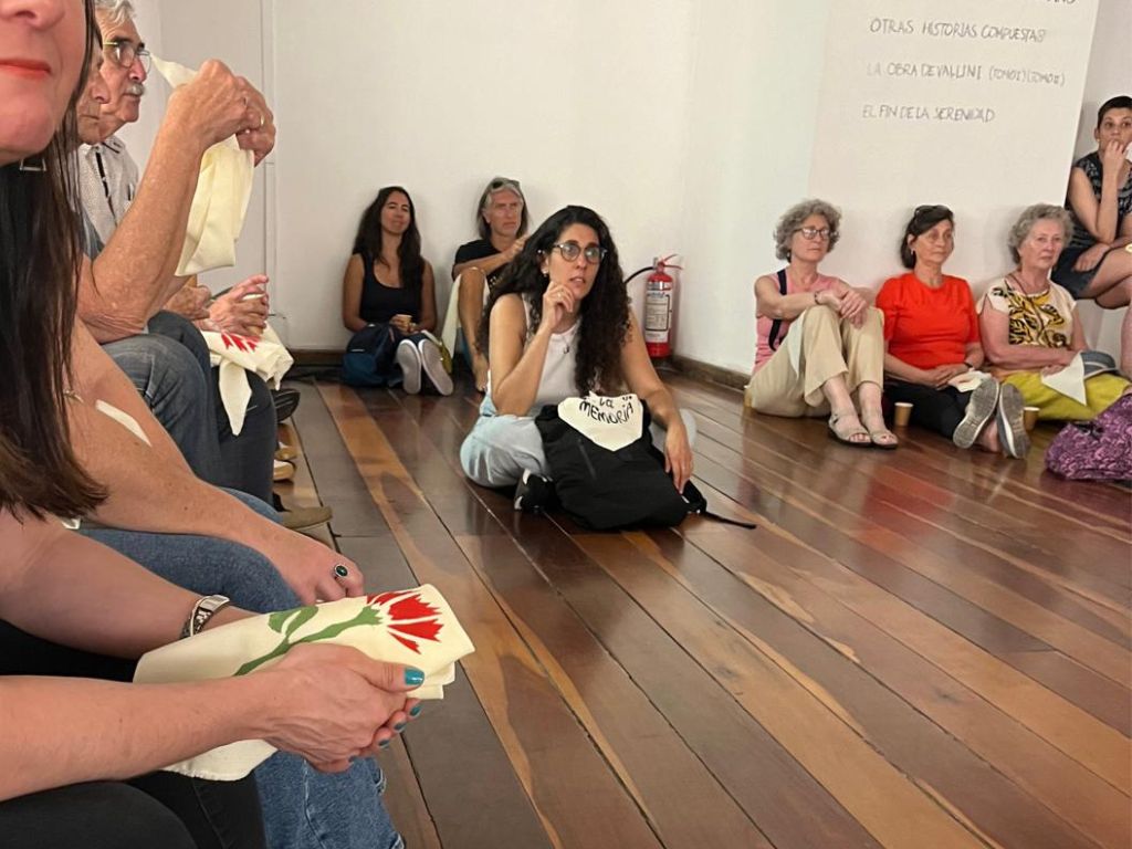 Fotografía con varias personas sentadas sobre un piso de madera, con su atención dirigida al frente. Algunos tienen un pañuelo en sus manos con un clavel rojo