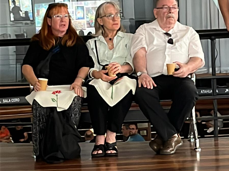 Fotografía de tres personas sentadas en la que se puede ver que dos de ellas tienen sobre sus piernas un pañuelo triangular con la imagen de un clavel rojo