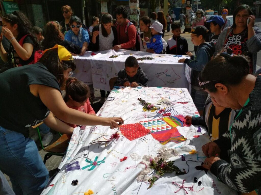 Fotografía tomada en la vía pública en la que se pueden ver personas de todas las edades, participando del bordado del mantel, el cual se encuentra extendido. sobre dos mesas. Al fondo de la fotografía se observa transito y otras personas circulando por la vereda.