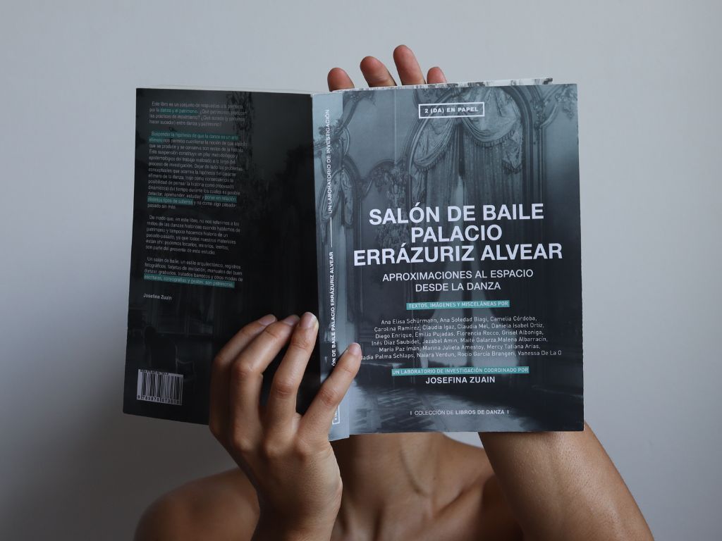 Fotografía de fondo gris con la imagen del torso de una persona que sostiene un libro abierto que le cubre el rostro.