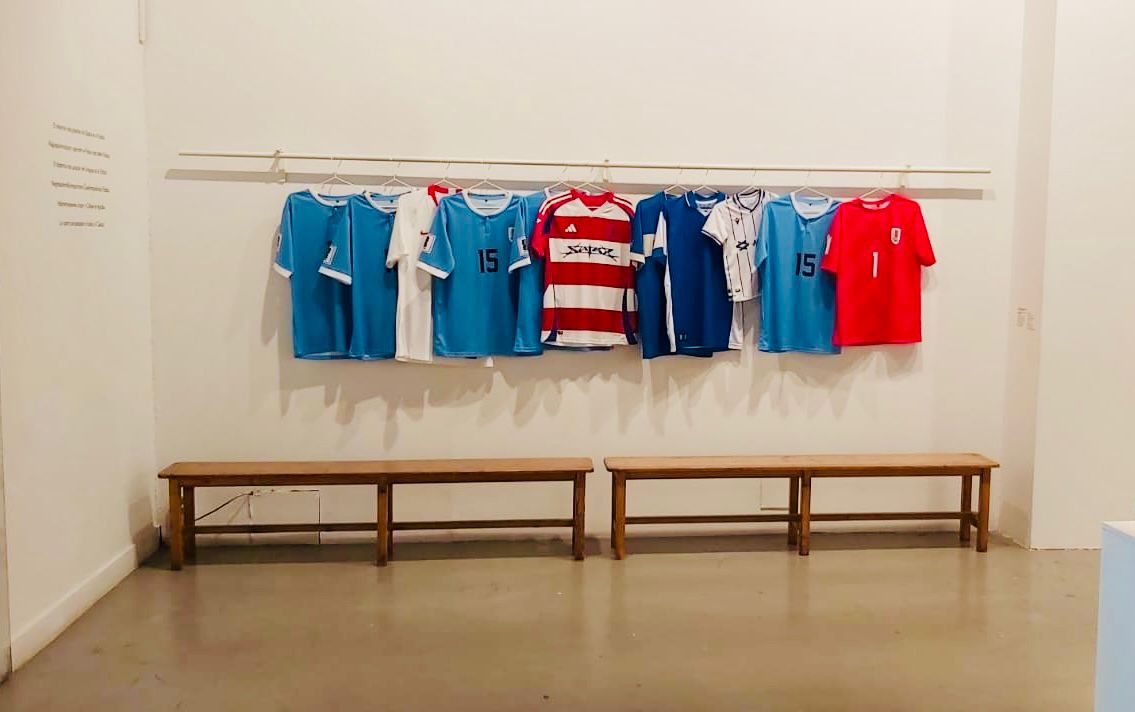 Imagen de la muestra donde se ven diferentes camisetas de fútbol de selecciones colgadas en lo que parece un vestuario, debajo de ellas se ven dos bancos largos.