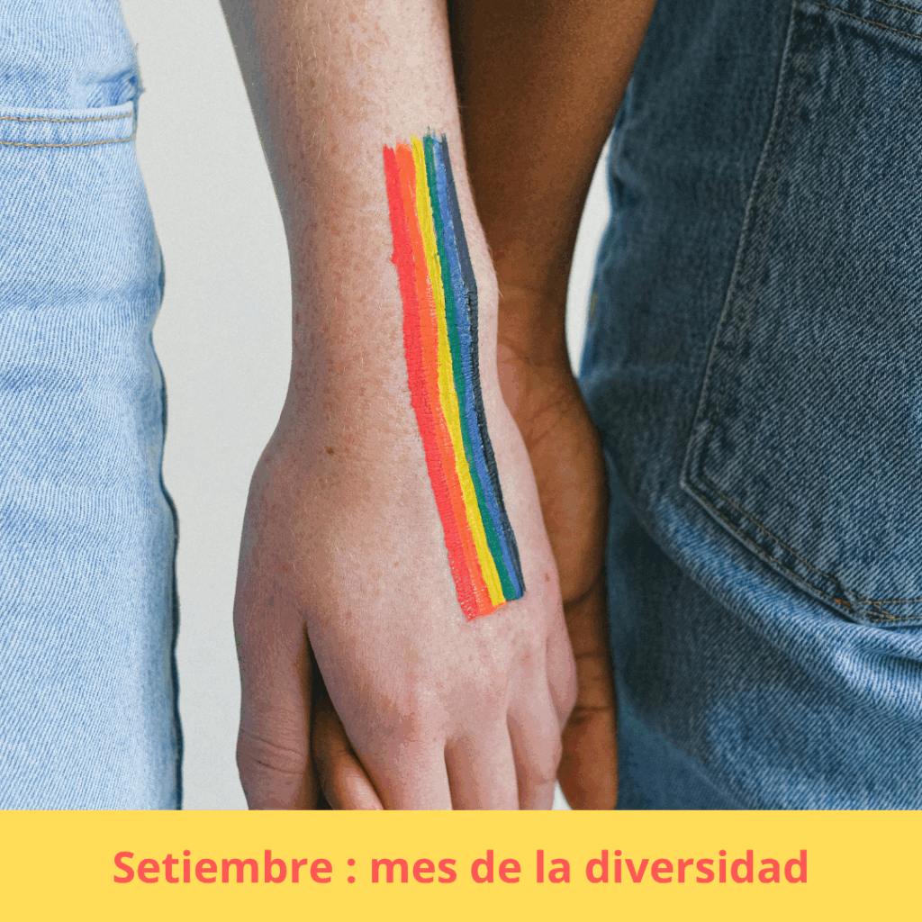 Imagen alusiva al mes de la diversidad. Primer plano de dos personas tomadas de la mano. Sobre la mano más visible hay una bandera del orgullo pintada con franjas verticales de colores. 