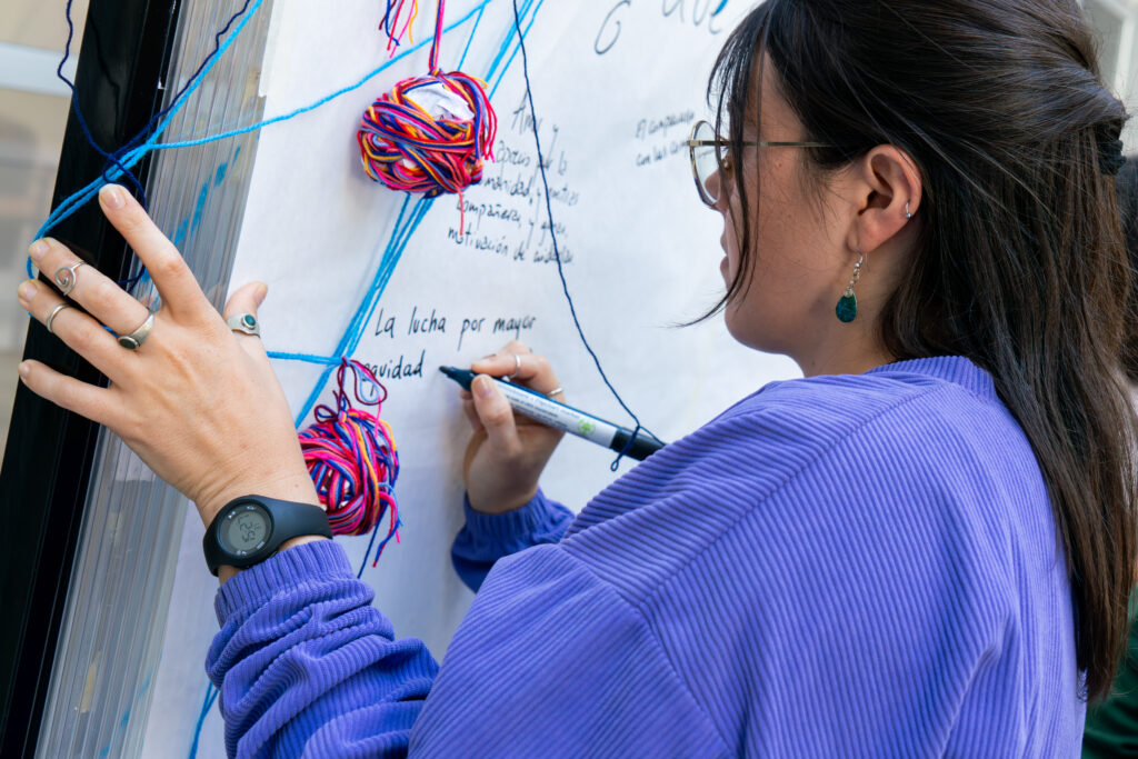 Imagen de mujer de perfil frente a un papelógrafo o cartelera, donde está escribiendo. Se ven diferentes hilos y texturas que atraviesan la cartelera